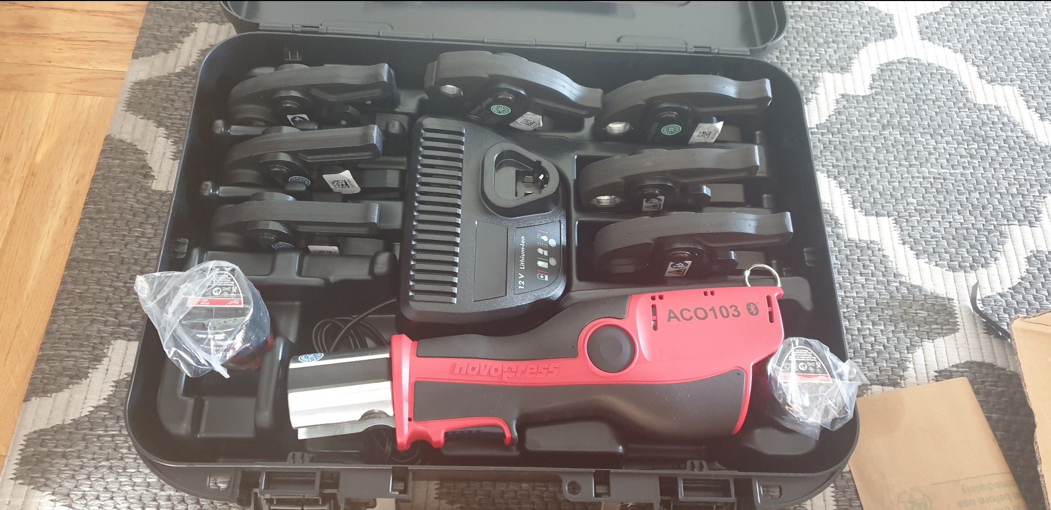 Zestaw narzędzi do zaciskania rur w czarnej walizce, widoczna zaciskarka Novopress ACO103, szczęki zaciskowe, ładowarka do akumulatora, całość na tle dywanu w geometryczne wzory.