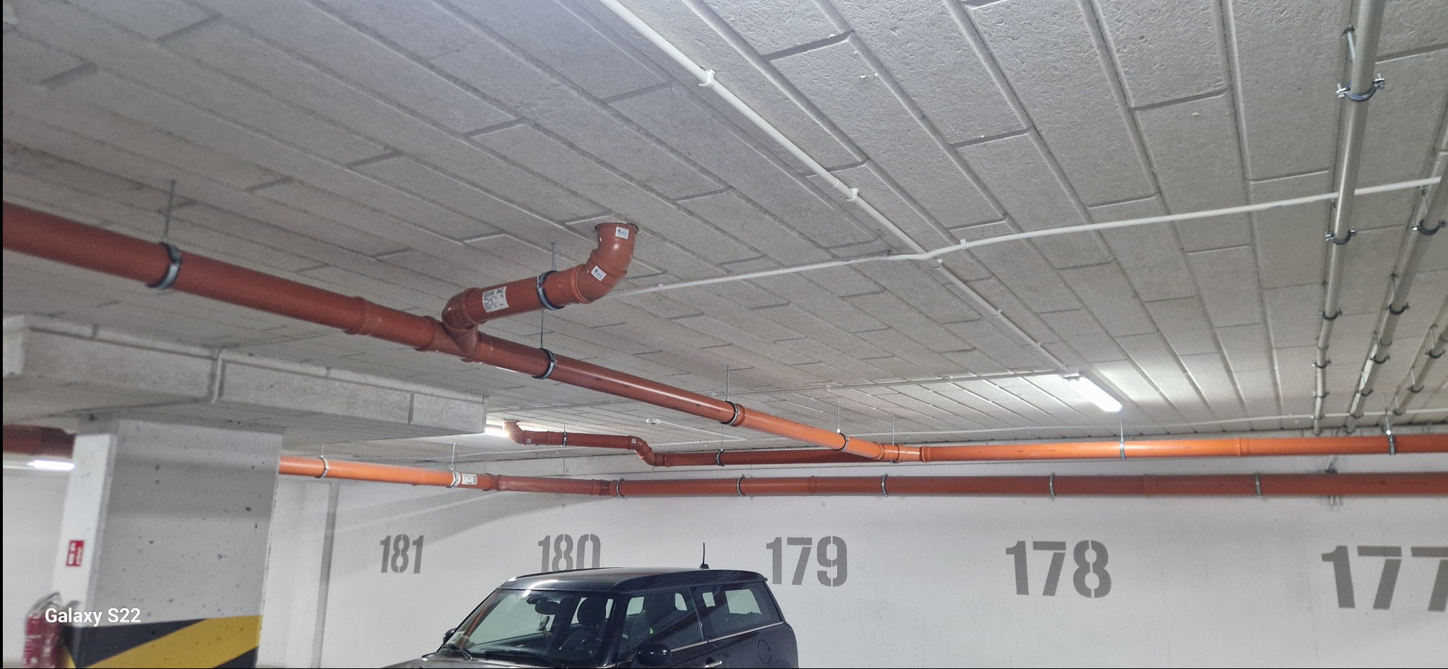 Pomarańczowe rury kanalizacyjne pod sufitem parkingu podziemnego, widoczne numery miejsc parkingowych na ścianie.