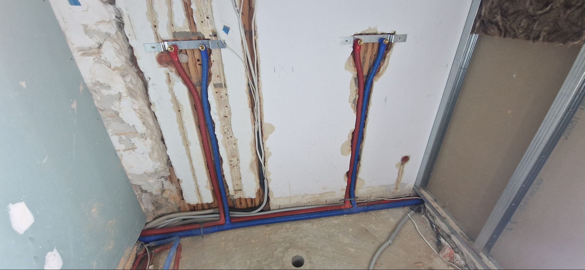 Instalacja wodna z rur PEX w kolorze czerwonym i niebieskim, mocowanych do ściany za pomocą metalowych uchwytów, widoczne przewody elektryczne i fragmenty niezabezpieczonej ściany z ubytkami tynku.