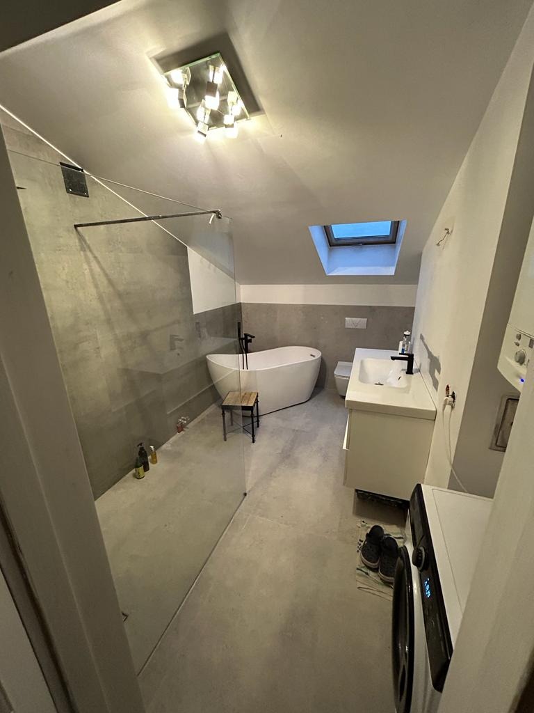 Nowoczesna łazienka na poddaszu z przeszkloną kabiną prysznicową, wolnostojącą wanną, białą umywalką i pralką, oświetlona lampą sufitową i światłem z okna dachowego.