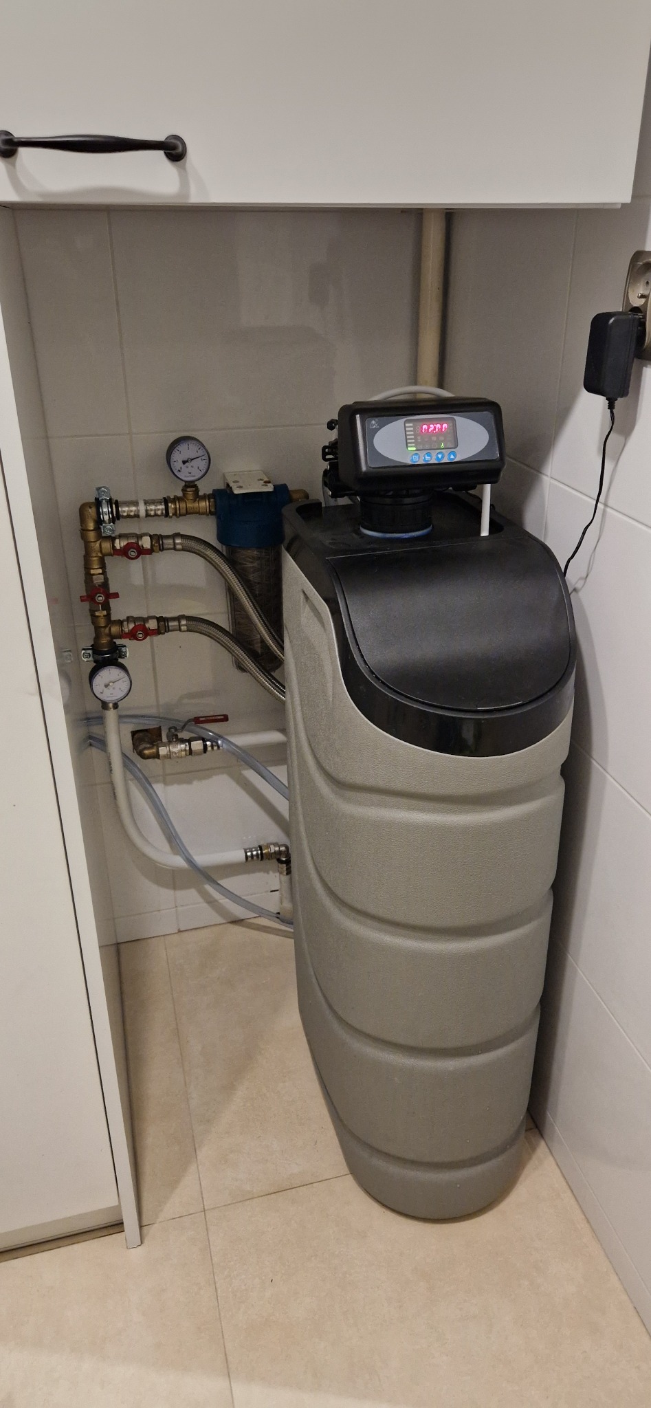 Szaro-czarna stacja uzdatniania wody z cyfrowym wyświetlaczem, podłączona do instalacji wodnej w pomieszczeniu z białymi płytkami i szafką.
