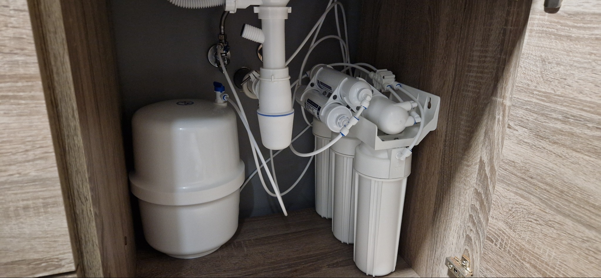Biały system filtracji wody osmotycznej zamontowany w szafce pod zlewem, widoczne rury doprowadzające wodę i zbiornik ciśnieniowy.