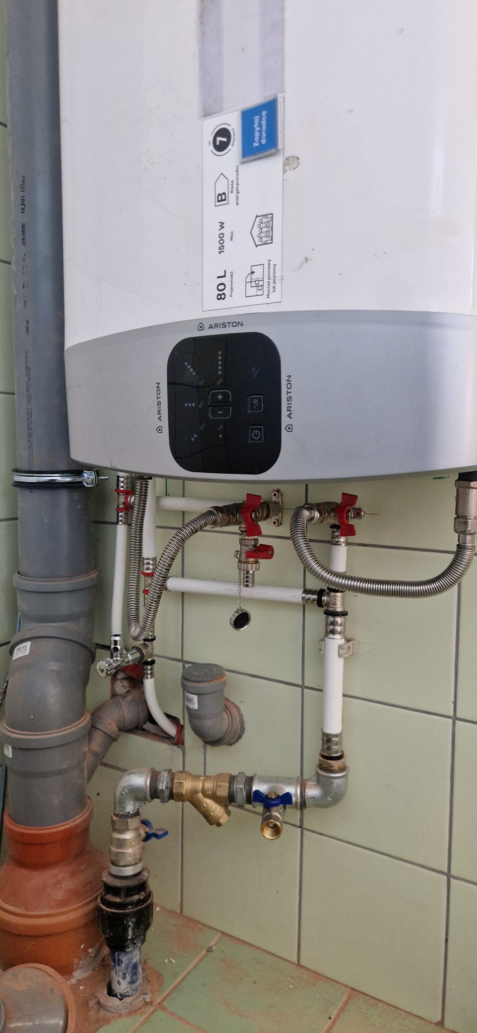 Bojler Ariston 80L z podłączonymi instalacjami hydraulicznymi w łazience z zielonymi płytkami, widoczne liczne rury z zaworami, zasilanie i odpływ.