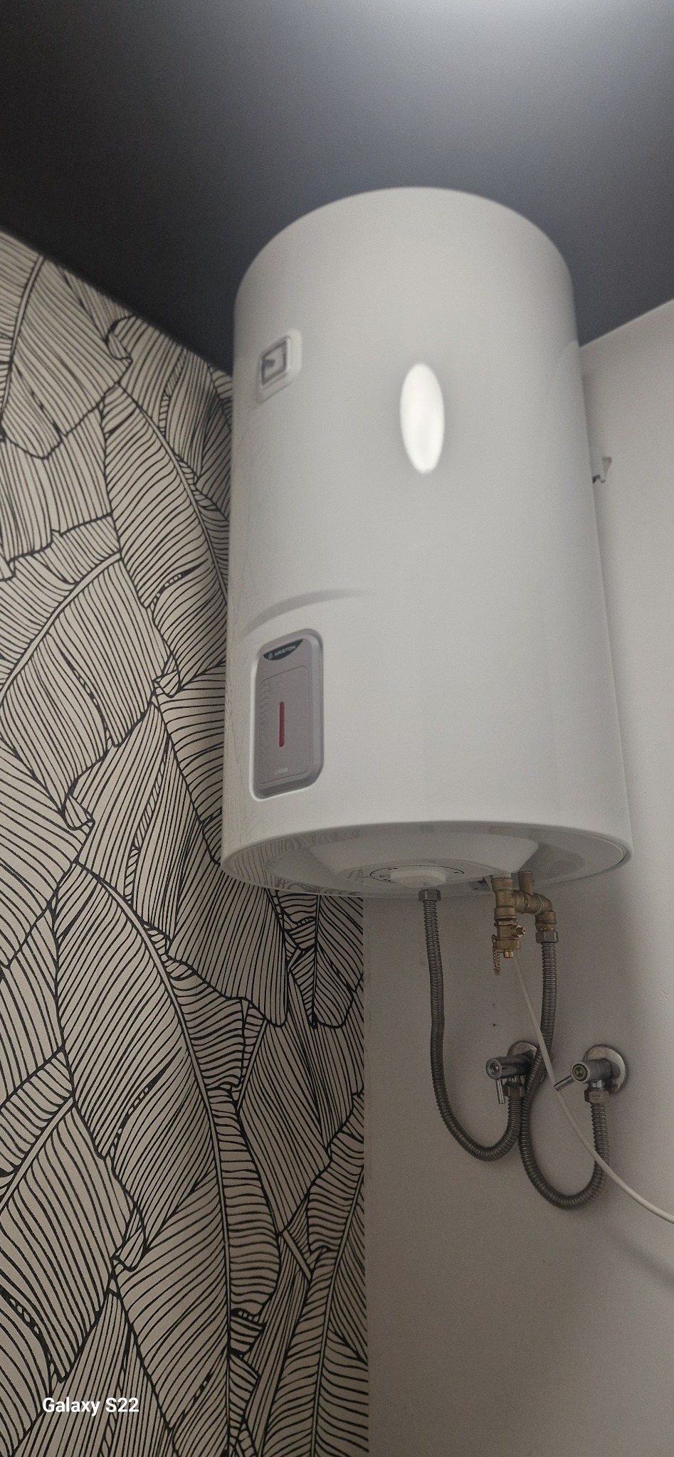 Bojler elektryczny Ariston zamontowany na ścianie z tapetą w liście, widoczne przyłącza wodne i zawory.