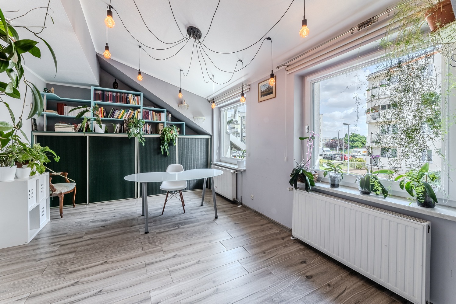 Jasne wnętrze poddasza z ukośnym sufitem i oknami, wyposażone w biurko z krzesłem, regał z książkami i zielonymi roślinami, oświetlone wiszącymi żarówkami na kablach.