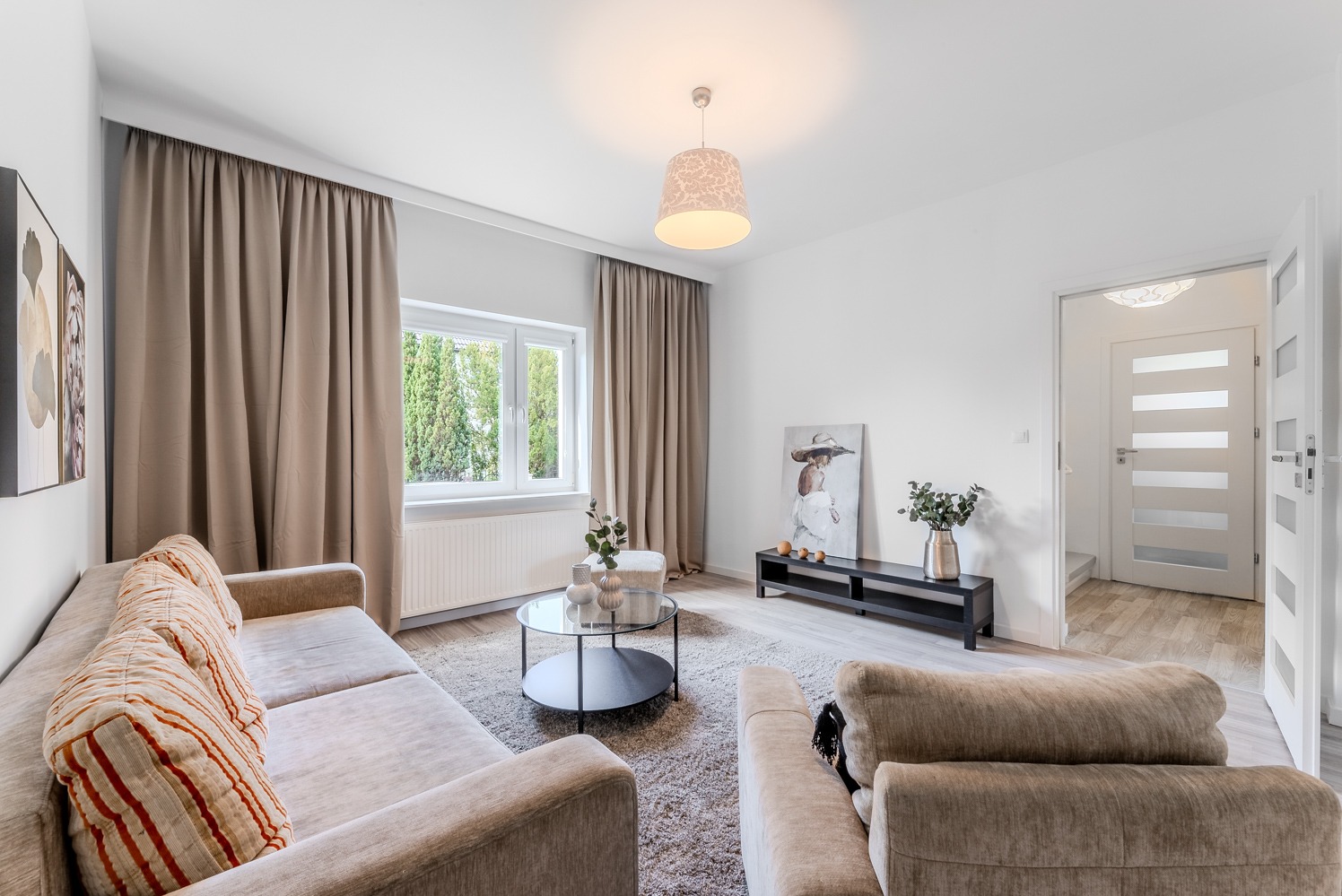 Jasny salon z beżową sofą w paski, okrągłym stolikiem kawowym z szklanym blatem, obrazem kobiety w kapeluszu i roślinami w wazonie, oświetlony lampą z abażurem w kwiaty.