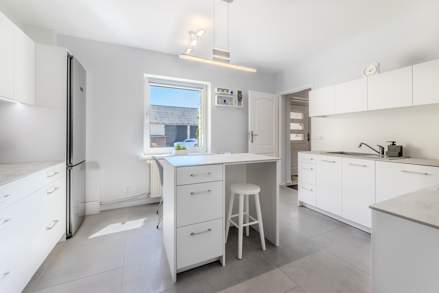 Jasna kuchnia z białymi meblami, wyspą kuchenną z szufladami i stołkiem, nowoczesnym oświetleniem i widokiem na podwórko przez okno.