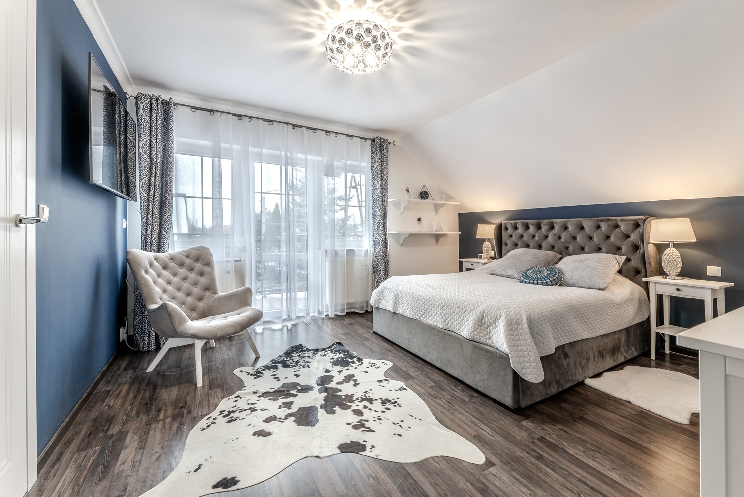 Jasna sypialnia z ciemnoszarą ścianą, łóżkiem z pikowanym zagłówkiem, wzorzystym dywanem imitującym skórę krowy i fotelem typu uszak w kolorze beżowym