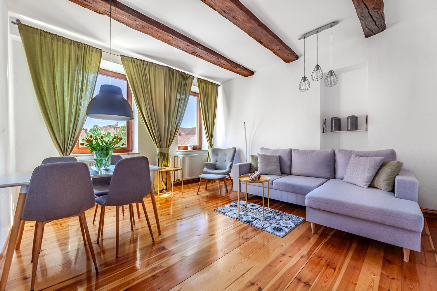 Jasne wnętrze z drewnianą podłogą, belkami stropowymi, szarym narożnikiem, stołem z krzesłami i zielonymi zasłonami.