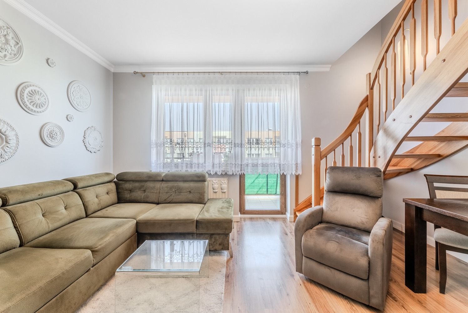 Salon z narożną sofą w odcieniach zieleni, szklanym stolikiem kawowym, ozdobnymi rozetami na ścianie, drewnianymi schodami i fotelem typu relaks.