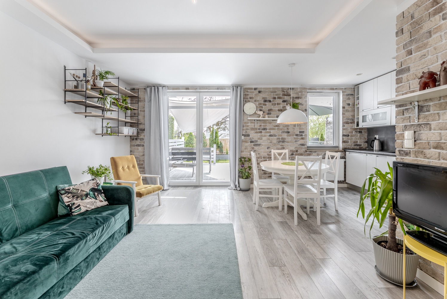 Jasne wnętrze z ceglaną ścianą, zieloną sofą, białym stołem i krzesłami, oraz półkami z roślinami, widok na taras przez przesuwne drzwi.