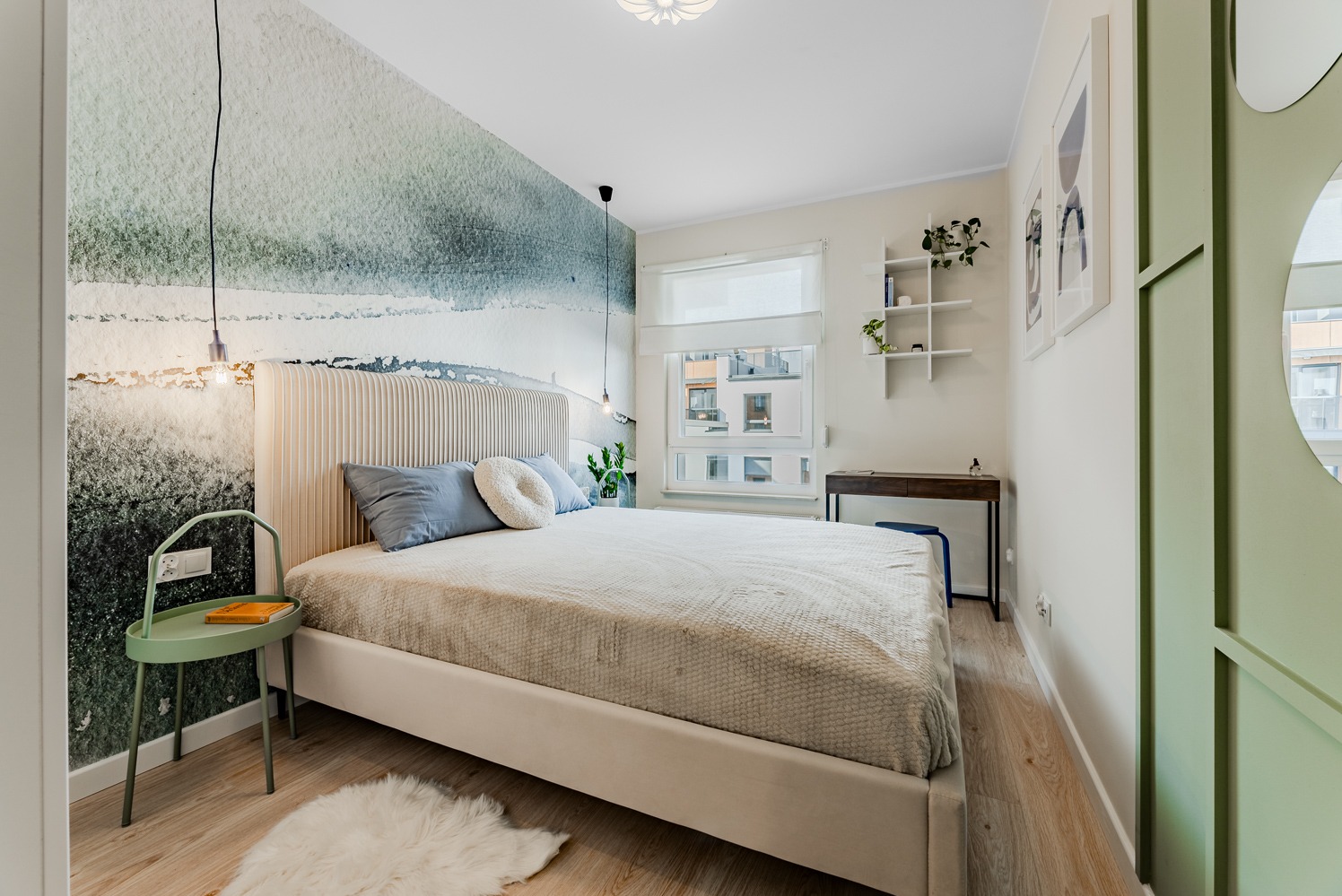 Jasna sypialnia z łóżkiem z wezgłowiem w prążki, akwarelową tapetą imitującą krajobraz i zielonym stolikiem nocnym z książką.