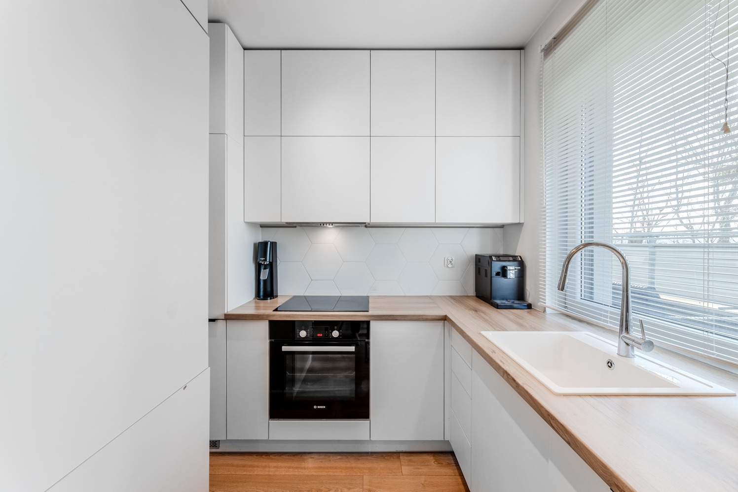 Minimalistyczna kuchnia z białymi szafkami, drewnianym blatem, heksagonalnymi płytkami nad blatem i widokiem z okna przysłoniętego żaluzjami.