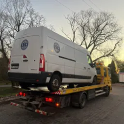 Żółta laweta z białym busem transportującym leki i ratującym życie, zaparkowana na brukowanym parkingu pod wieczornym niebem.