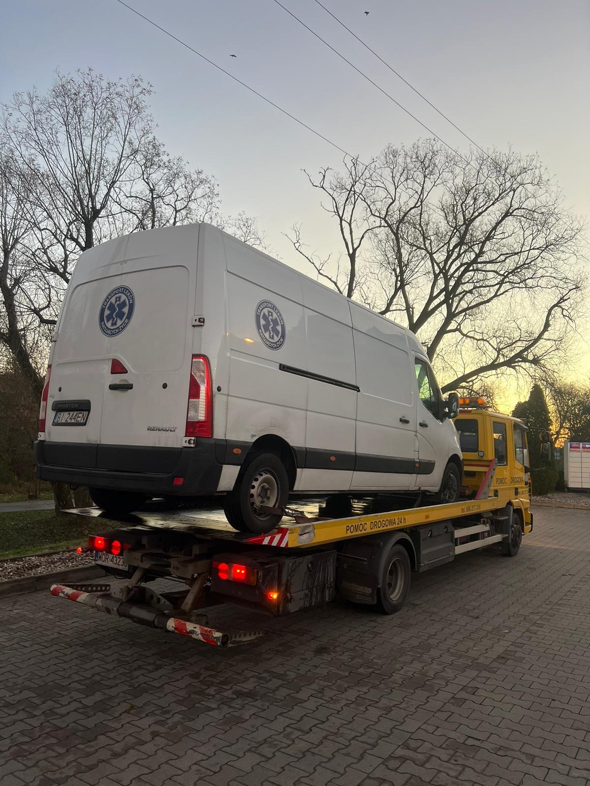 Żółta laweta z białym busem transportującym leki i ratującym życie, zaparkowana na brukowanym parkingu pod wieczornym niebem.