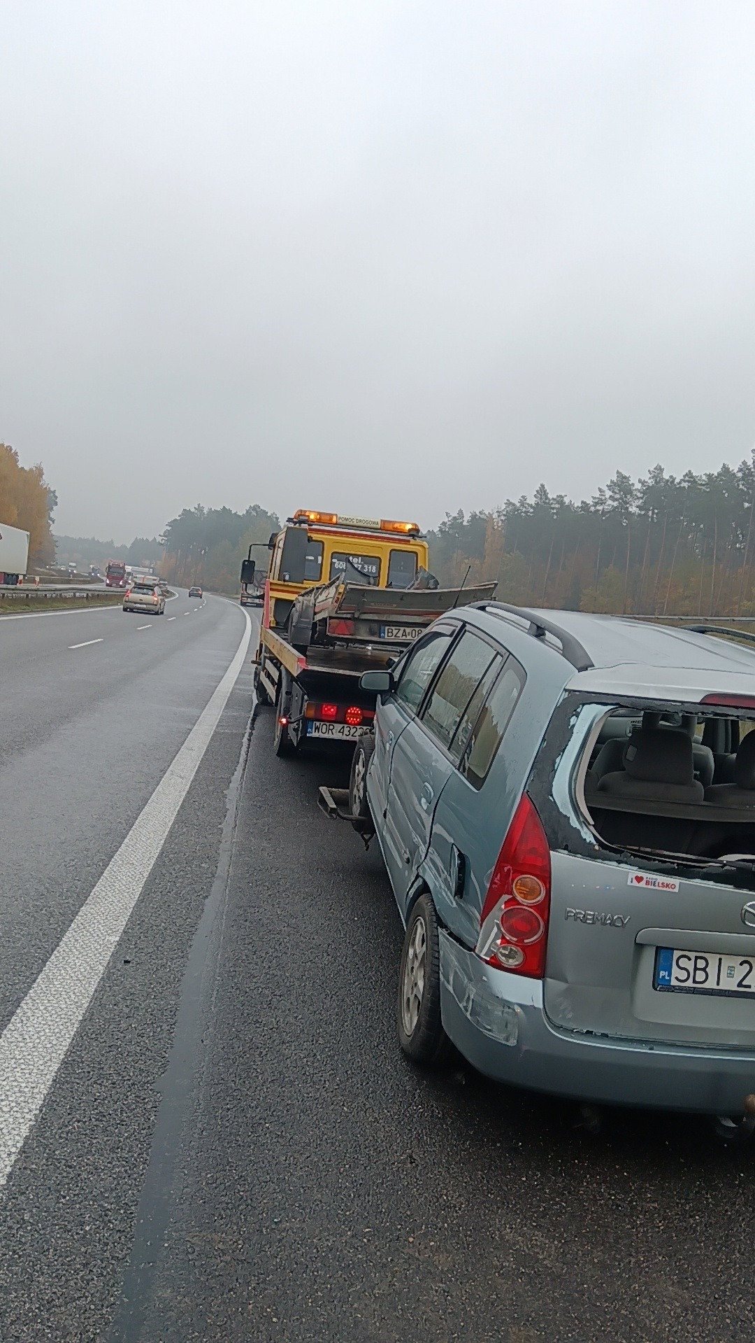 Uszkodzony, niebieski samochód Mazda Premacy z wybitą tylną szybą, holowany na autostradzie przez żółtą ciężarówkę pomocy drogowej z włączonymi światłami ostrzegawczymi; widoczna naklejka 'I love...