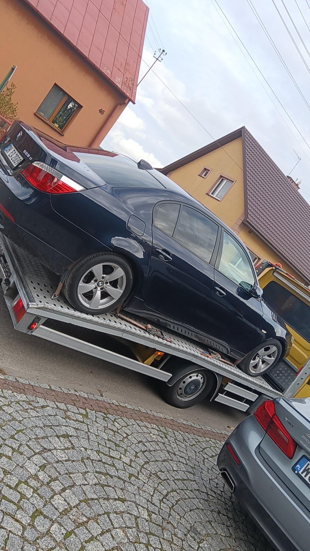 Czarny samochód BMW serii 5 (E60) zabezpieczony pasami na platformie lawety, widok z boku na tle budynków mieszkalnych.