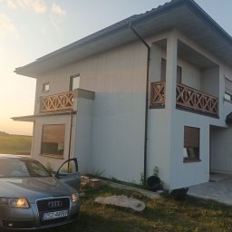 Sławomir Zieliński Konstrukcje drewniane - Nowoczesny dom z jasną elewacją i drewnianymi balustradami balkonowymi w kształcie krzyży, widok z zewnątrz, samochód osobowy z otwartymi drzwiami na pierwszym planie.