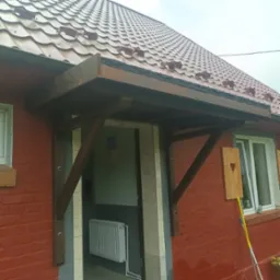 Zadaszenie wejściowe z ciemnego drewna, wsparte na ukośnych belkach, nad wejściem do budynku z czerwonej cegły. Obok okno z drewnianą ozdobą w kształcie serca. W tle widoczny fragment dachu krytego...