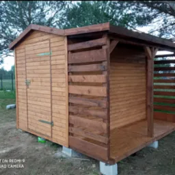 Drewniana konstrukcja z zamykaną częścią przypominającą domek narzędziowy i otwartą przestrzenią z drewnianą balustradą, całość posadowiona na betonowych bloczkach.