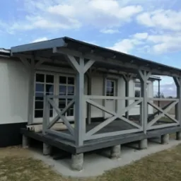 Zadaszony taras z drewnianą balustradą w kształcie krzyży, wsparty na betonowych słupach, przylegający do budynku z białymi ścianami i widocznymi rolkami materiału na podłodze.