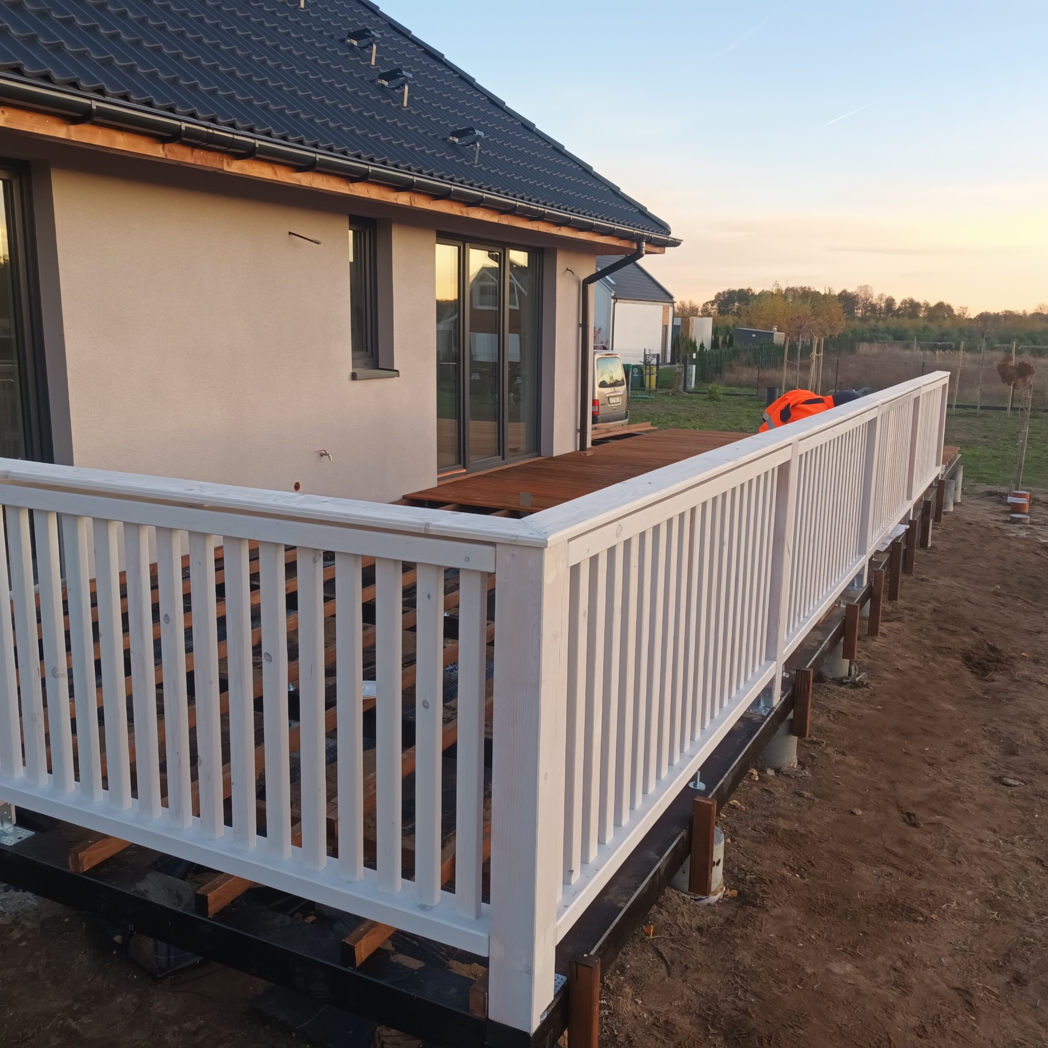 Wznoszenie białej, drewnianej balustrady na tarasie z ciemnego drewna, widok na dom jednorodzinny w tle podczas zachodu słońca.
