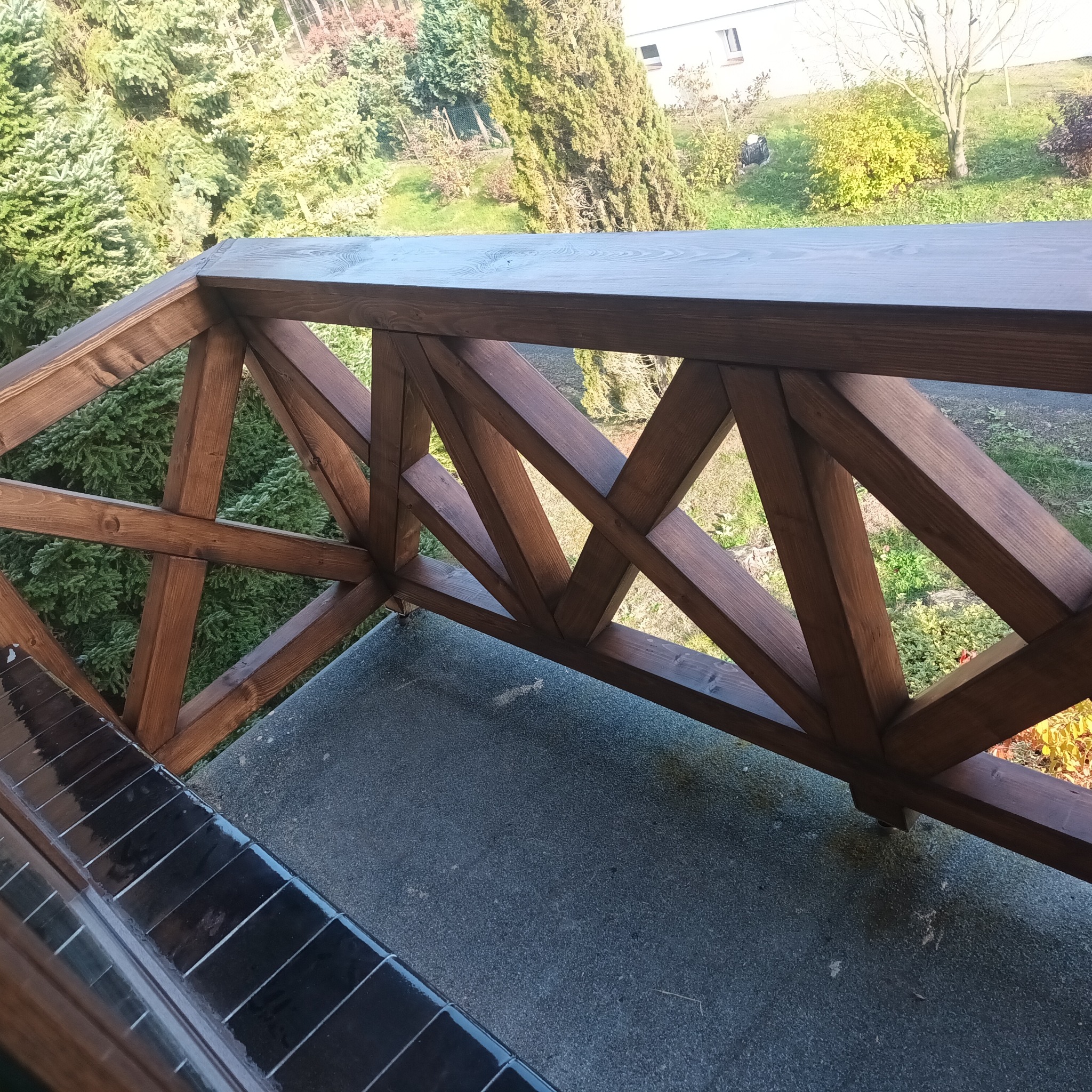 Drewniana balustrada balkonowa z krzyżowymi elementami, pomalowana na ciemny brąz, z widokiem na zielony ogród i budynek w tle.