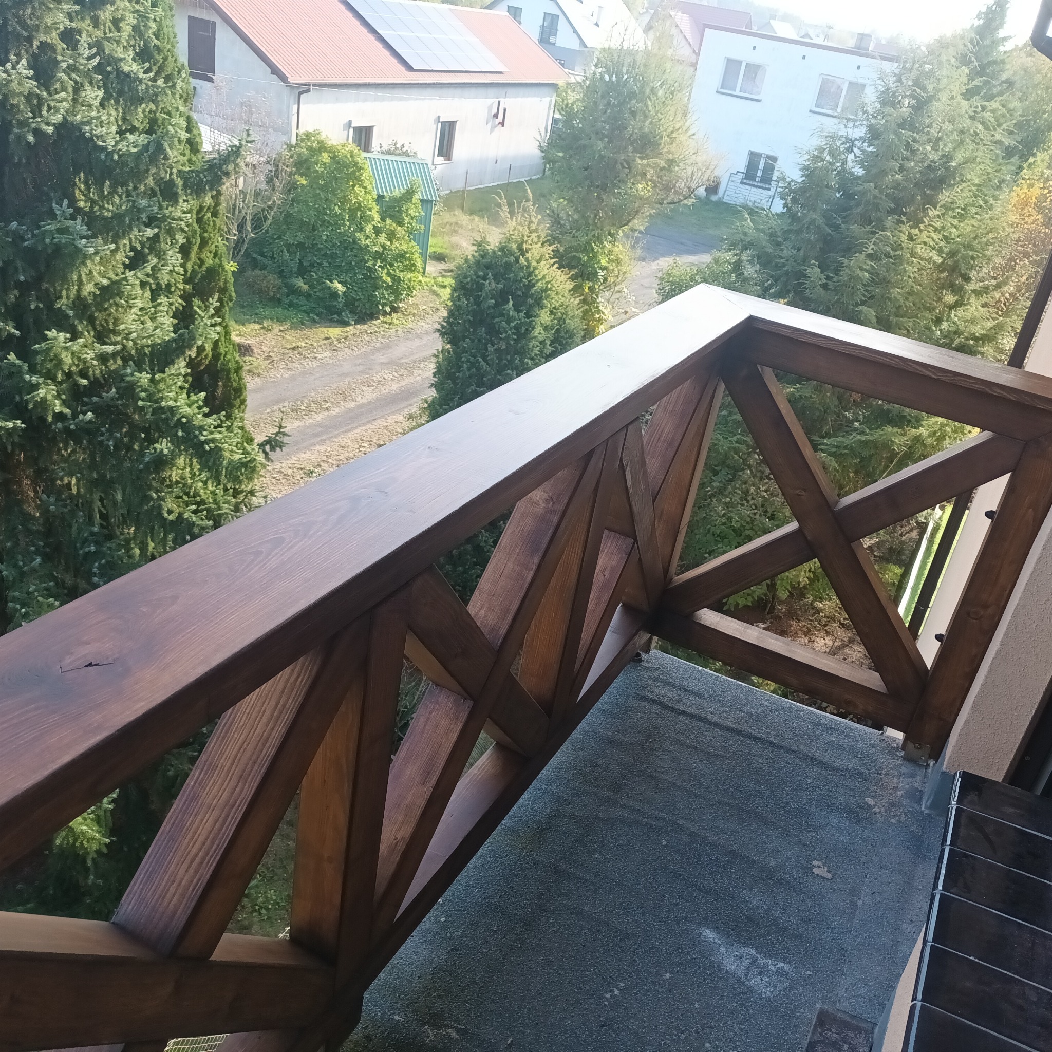 Ciemnobrązowa, drewniana balustrada balkonowa z geometrycznym wzorem krzyżujących się desek, widok z góry na otoczenie z domami i drzewami.