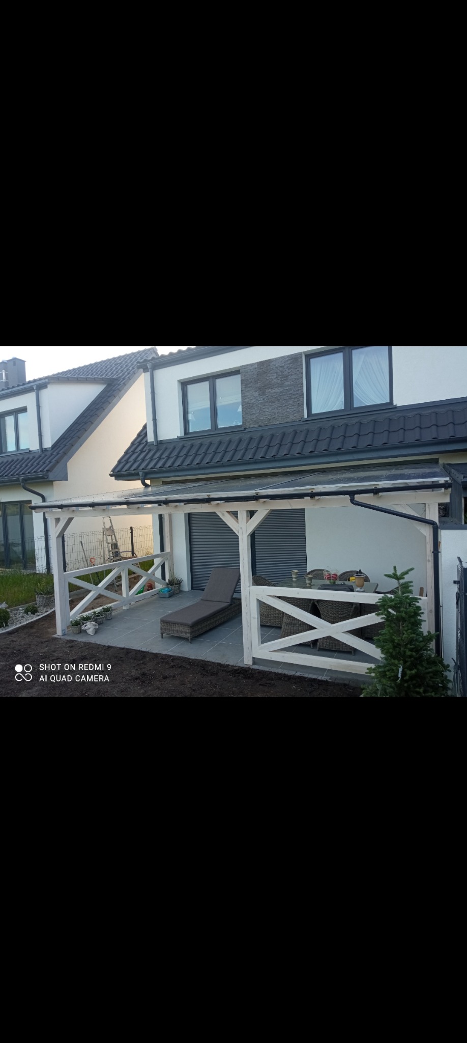 Nowoczesna, biała konstrukcja z drewna z zadaszeniem przylegająca do domu, z widocznym tarasem, leżakiem i meblami ogrodowymi. Widoczne świeżo posadzone rośliny.