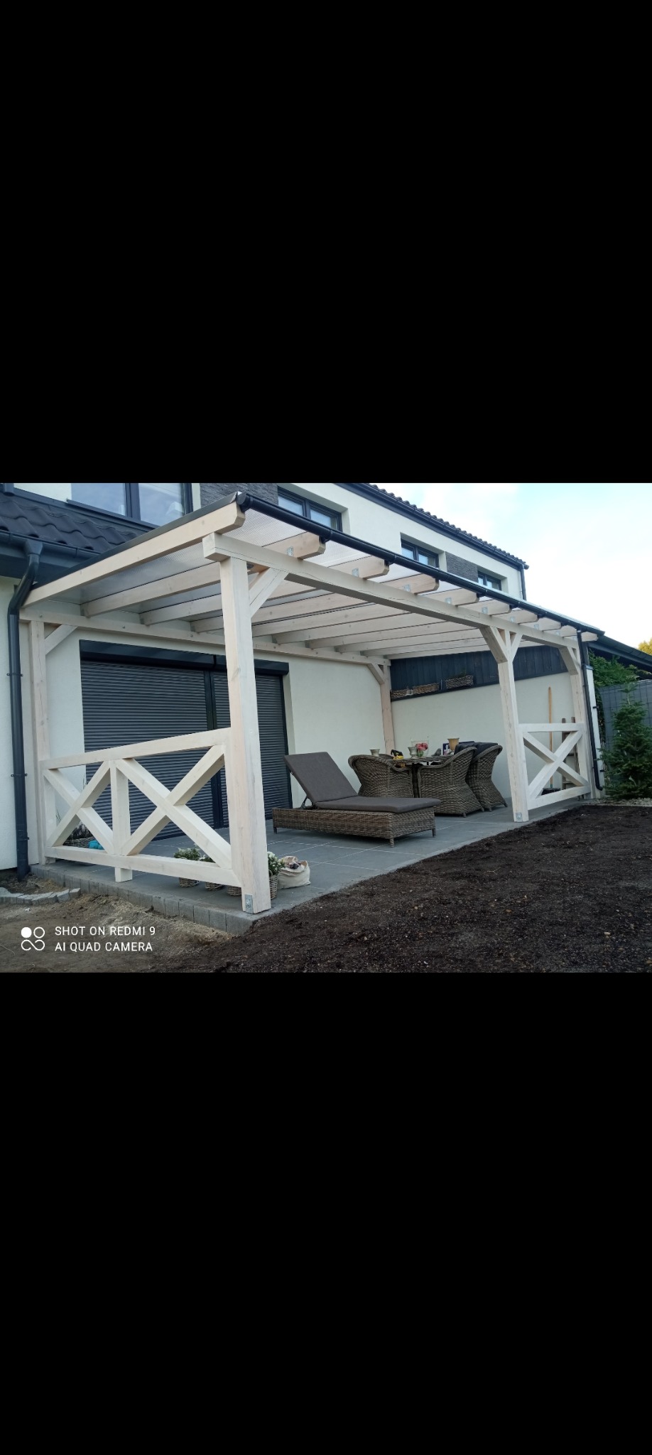 Biała drewniana pergola z ażurową balustradą, zadaszeniem z poliwęglanu, zestawem mebli ogrodowych i leżakiem, na wybrukowanym tarasie przylegającym do domu.