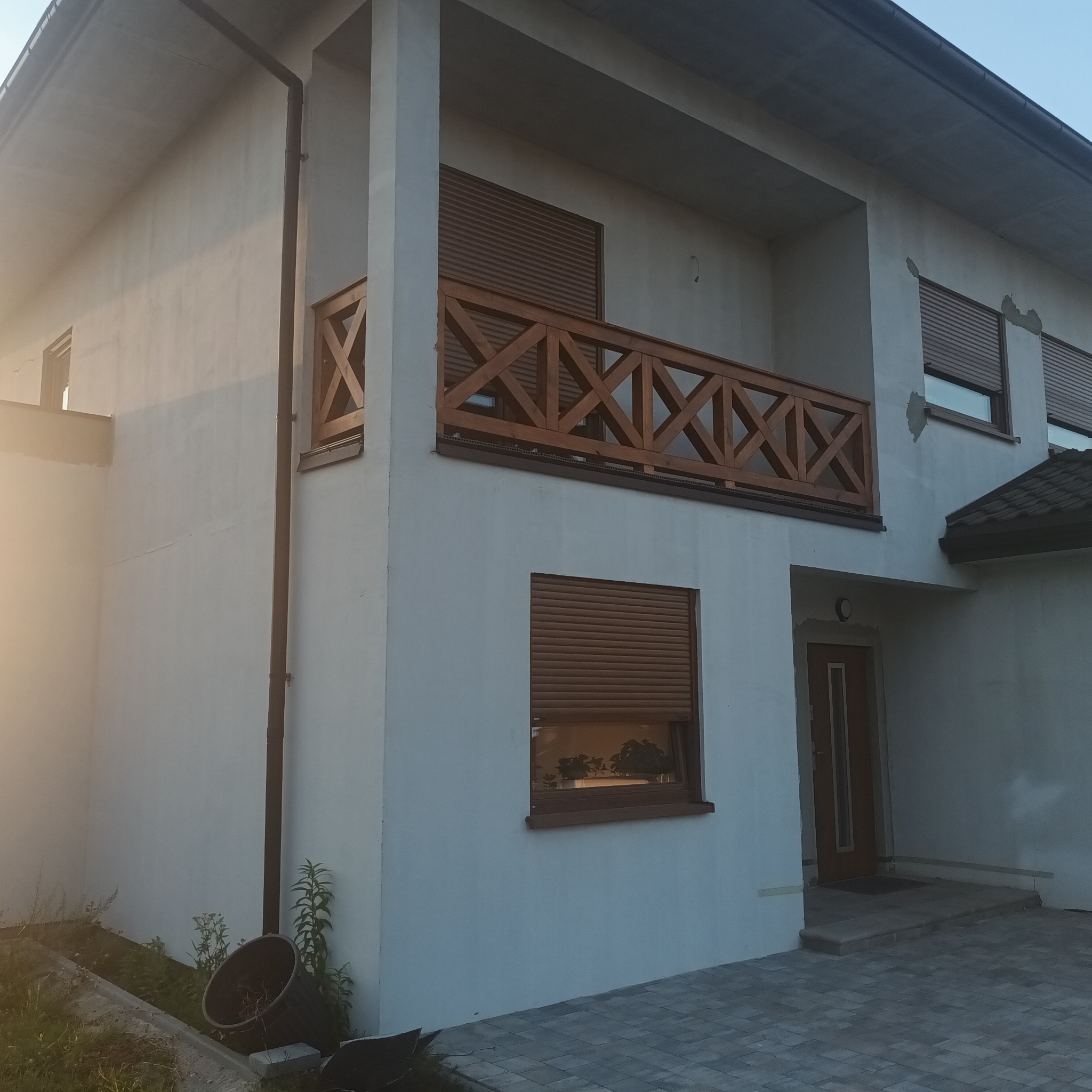 Dwupiętrowy dom z białymi ścianami, drewnianą balustradą balkonową w kratkę, brązowymi roletami w oknach i kostką brukową na podjeździe.