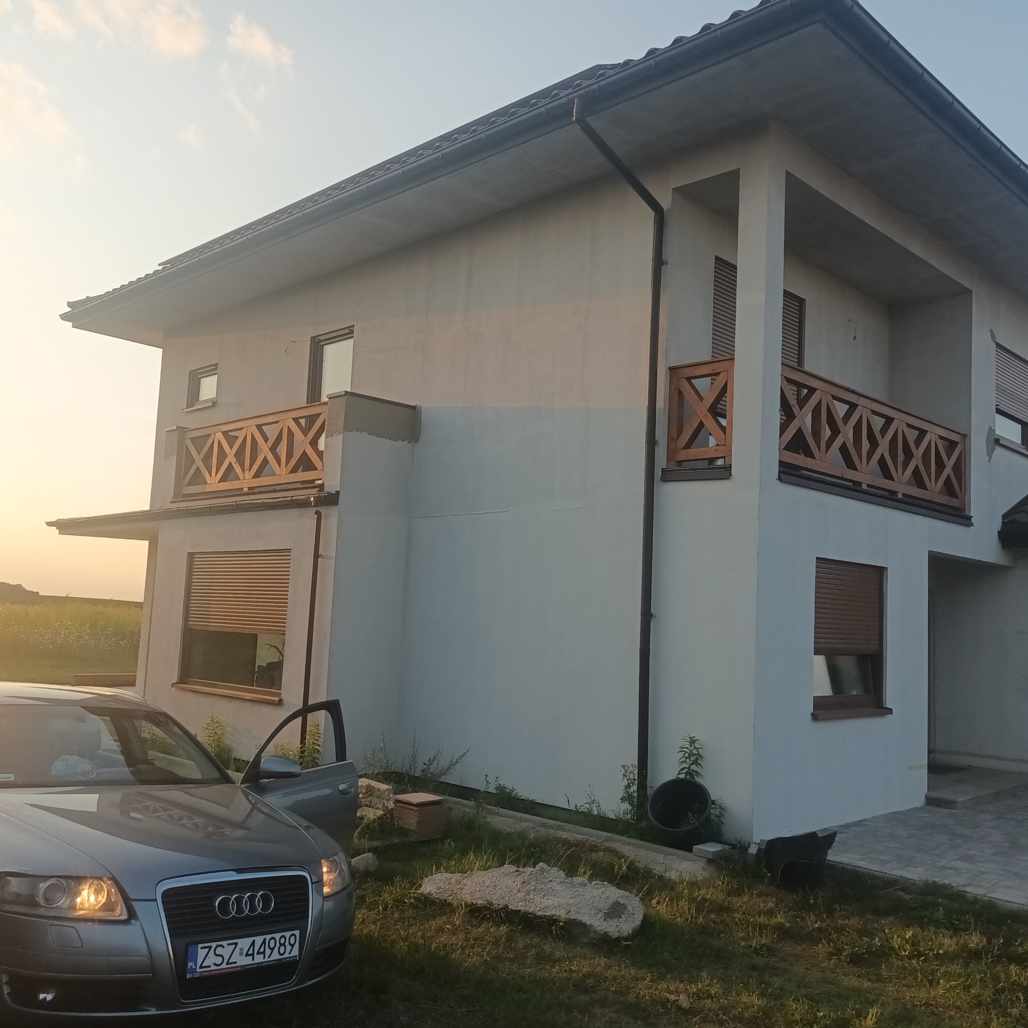 Nowoczesny dom z jasną elewacją i drewnianymi balustradami balkonowymi w kształcie krzyży, widok z zewnątrz, samochód osobowy z otwartymi drzwiami na pierwszym planie.