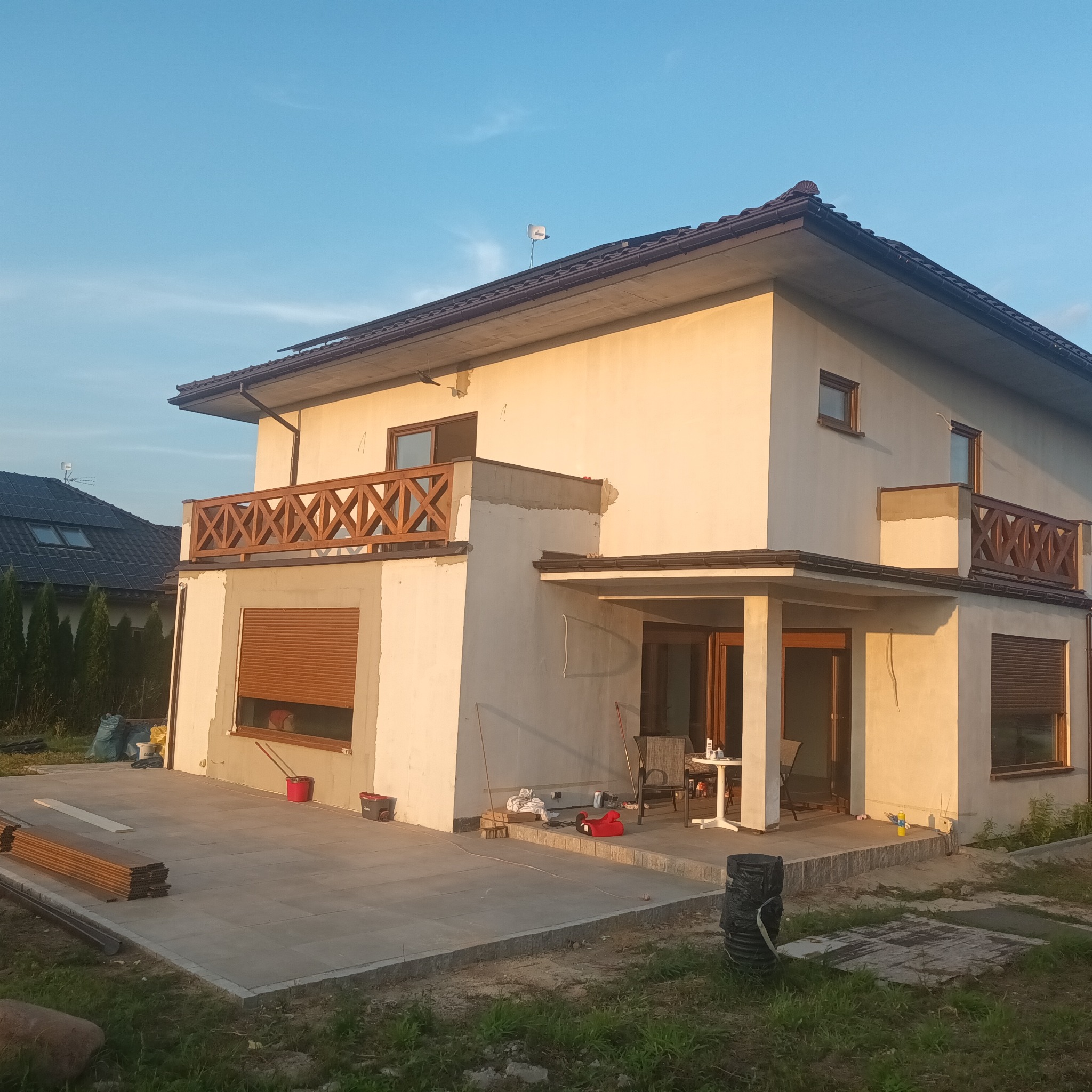 Dom w trakcie wykończenia z drewnianą balustradą na balkonie i nowym tarasem z płyt betonowych, widoczne materiały budowlane i narzędzia.