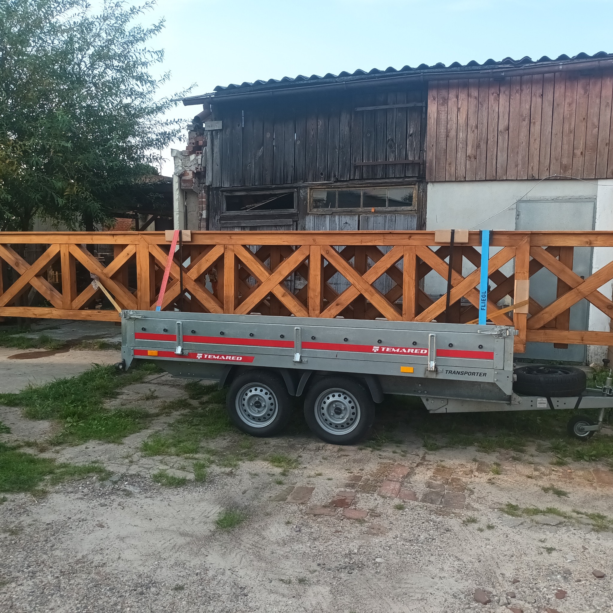 Element konstrukcyjny z drewna w kratkę, zabezpieczony pasami transportowymi na przyczepie samochodowej, na tle starego budynku gospodarczego.