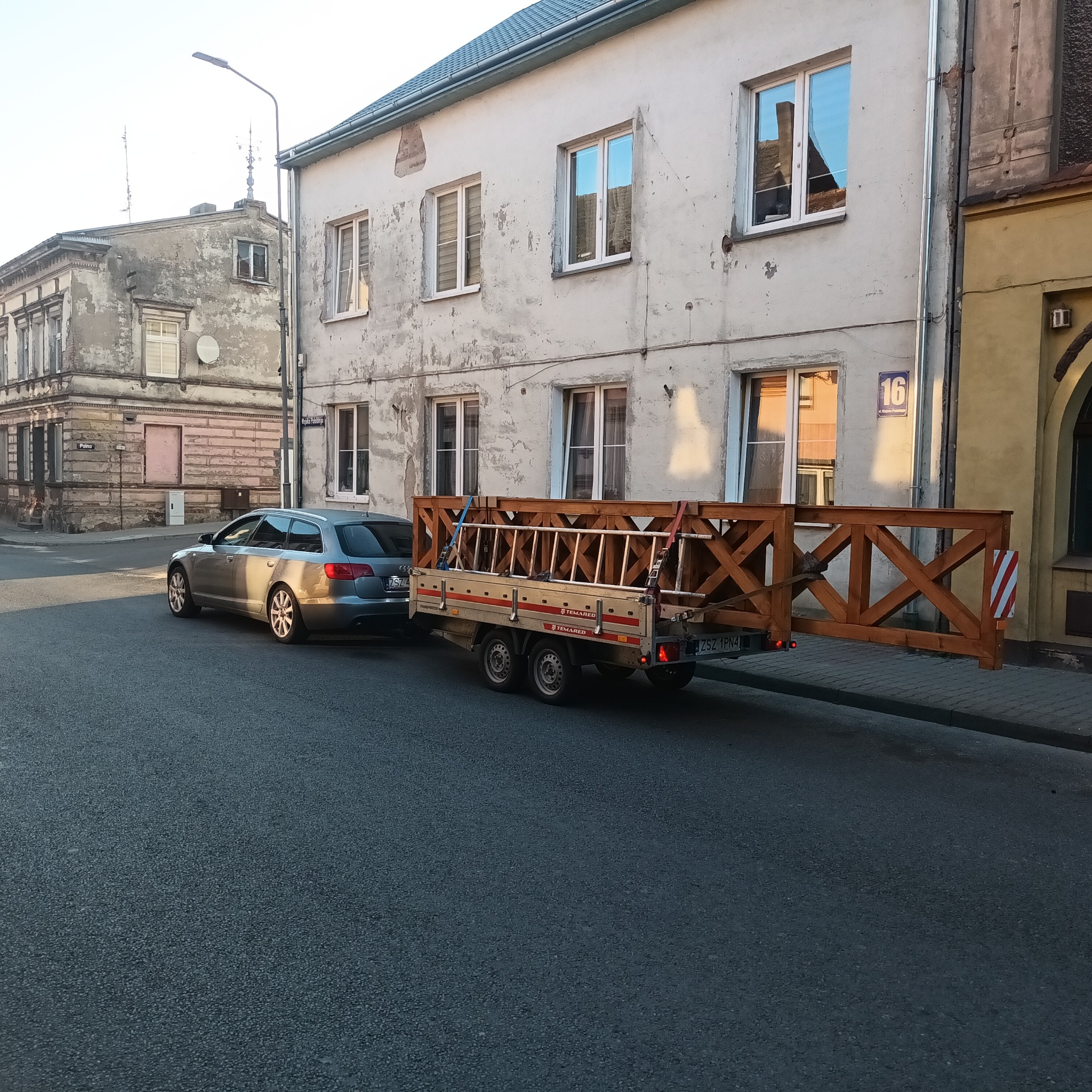 Fragment drewnianego ogrodzenia przewożony na przyczepie za samochodem osobowym na tle miejskiej zabudowy.