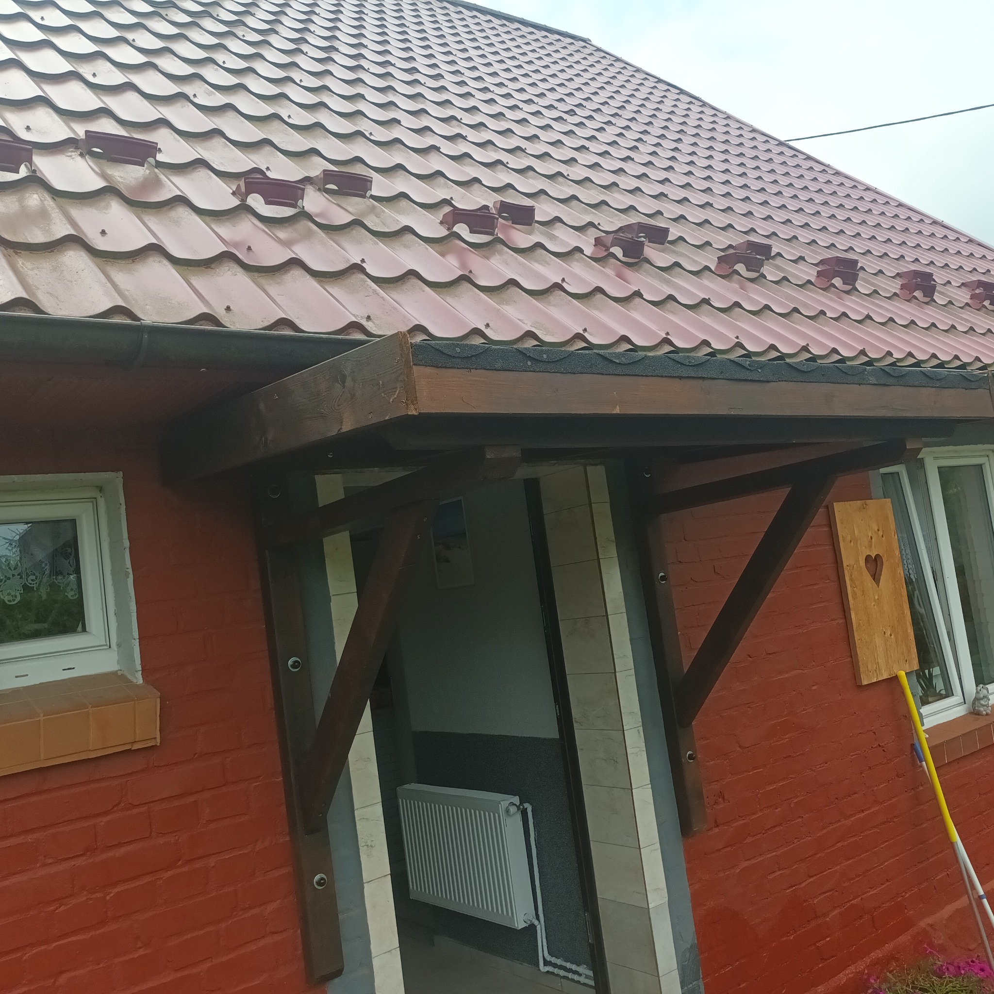 Zadaszenie wejścia do domu z drewnianą konstrukcją wspartą na dwóch słupach, czerwona ceglana ściana, dach z czerwonej blachy, widoczny fragment wnętrza z grzejnikiem.