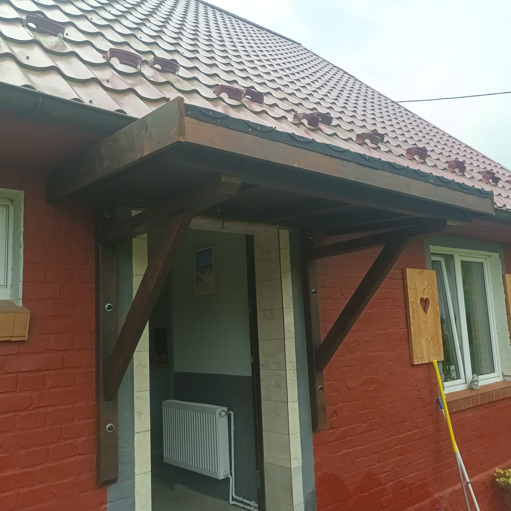 Zadaszenie wejściowe z ciemnego drewna, wsparte na ukośnych belkach, nad wejściem do budynku z czerwonej cegły. Obok okno z drewnianą ozdobą w kształcie serca. W tle widoczny fragment dachu krytego...