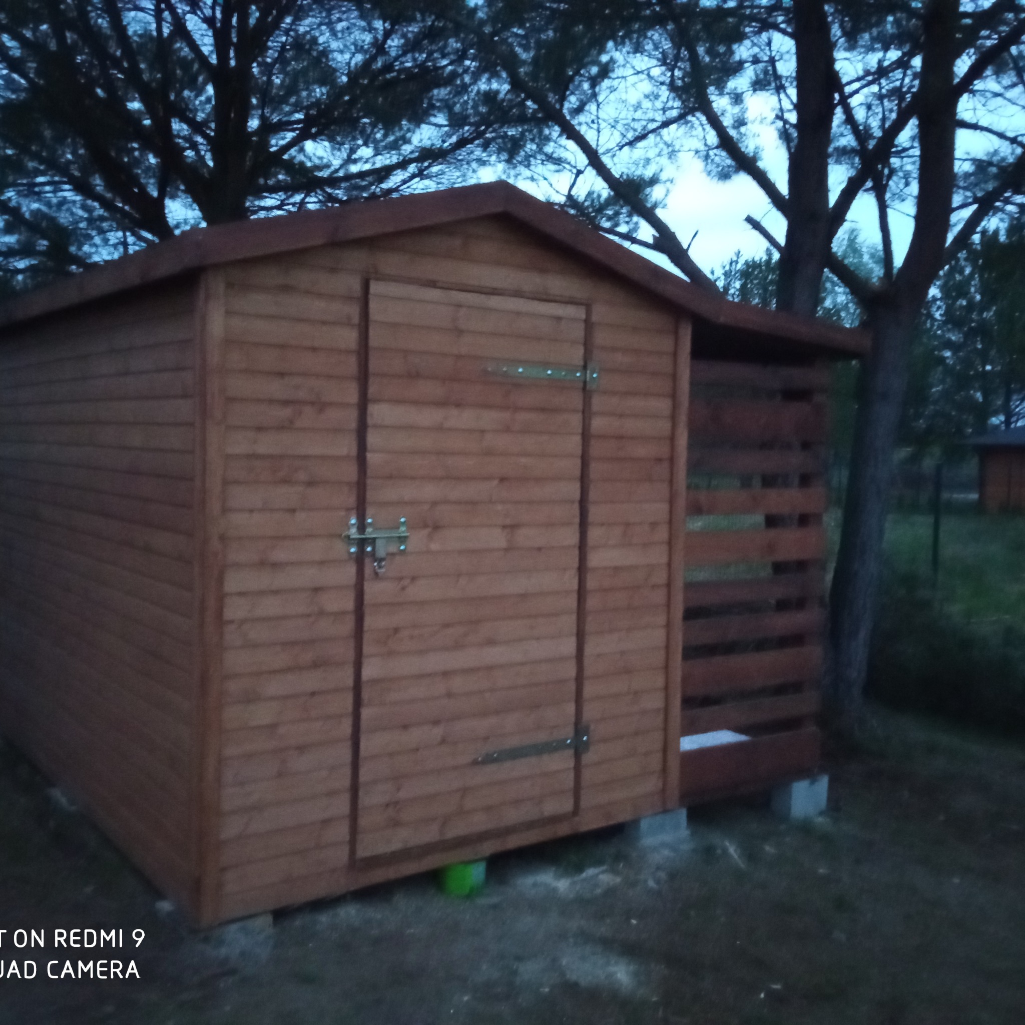 Drewniana budka ogrodowa z zamykanymi drzwiami i ażurową ścianką boczną, umieszczona na betonowych bloczkach, sfotografowana o zmierzchu na tle drzew.