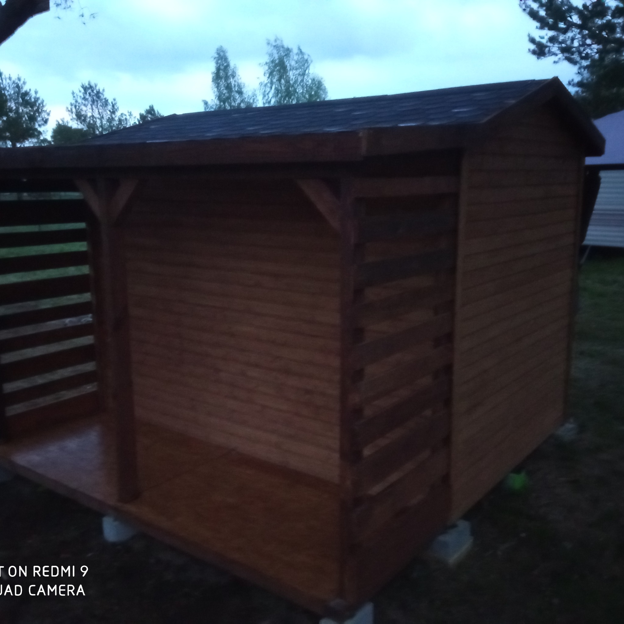 Drewniana konstrukcja z dachem krytym gontem bitumicznym, z ażurową ścianą boczną, posadowiona na betonowych bloczkach, sfotografowana o zmierzchu.