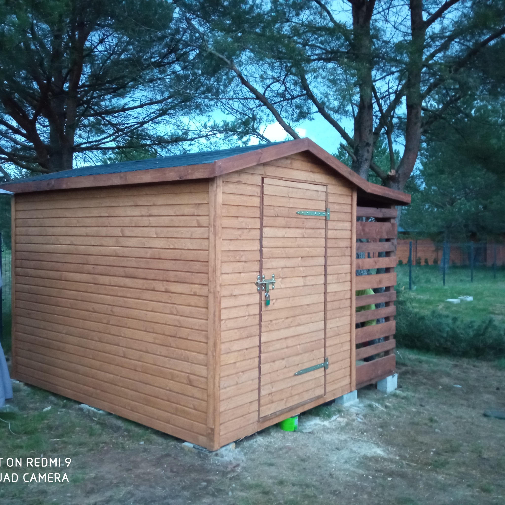 Drewniany domek narzędziowy z zamykanymi drzwiami i metalowymi zawiasami, umieszczony na betonowych bloczkach, z ażurową ścianką z boku, na tle trawnika i drzew