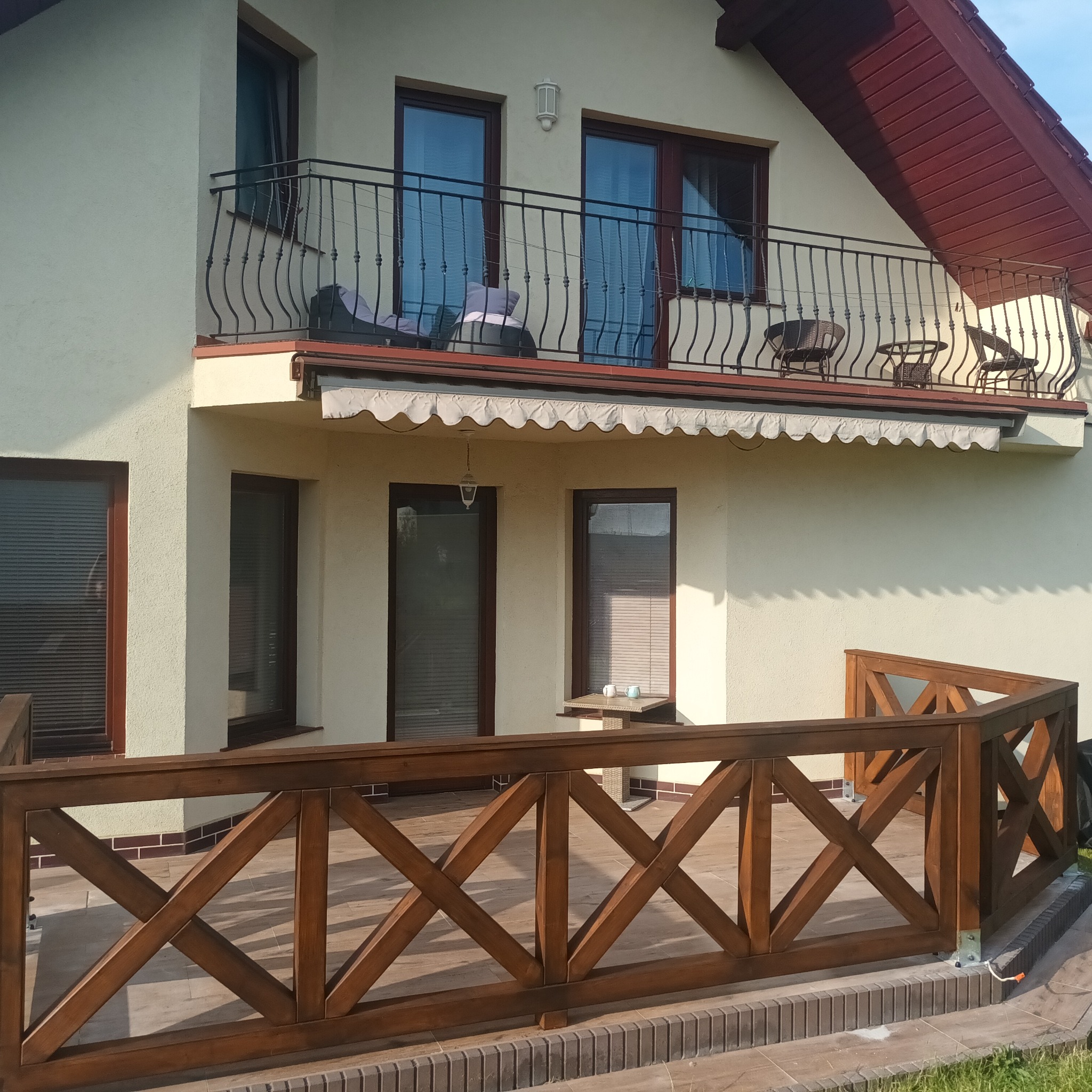 Zewnętrzny widok domu z drewnianym tarasem z balustradą o wzorze krzyżowym, balkon z kutą balustradą, markiza nad oknami i czerwony dach.