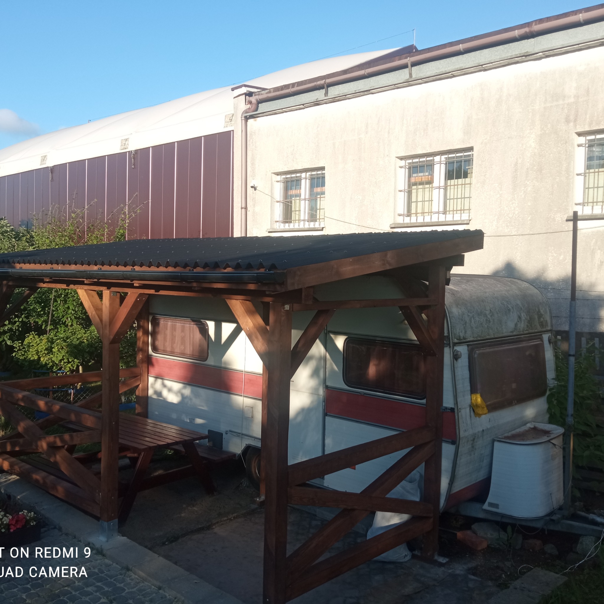 Drewniana konstrukcja zadaszona blachą falistą, z ławkami i stołem wewnątrz, stojąca obok starej przyczepy kempingowej na tle budynku gospodarczego.