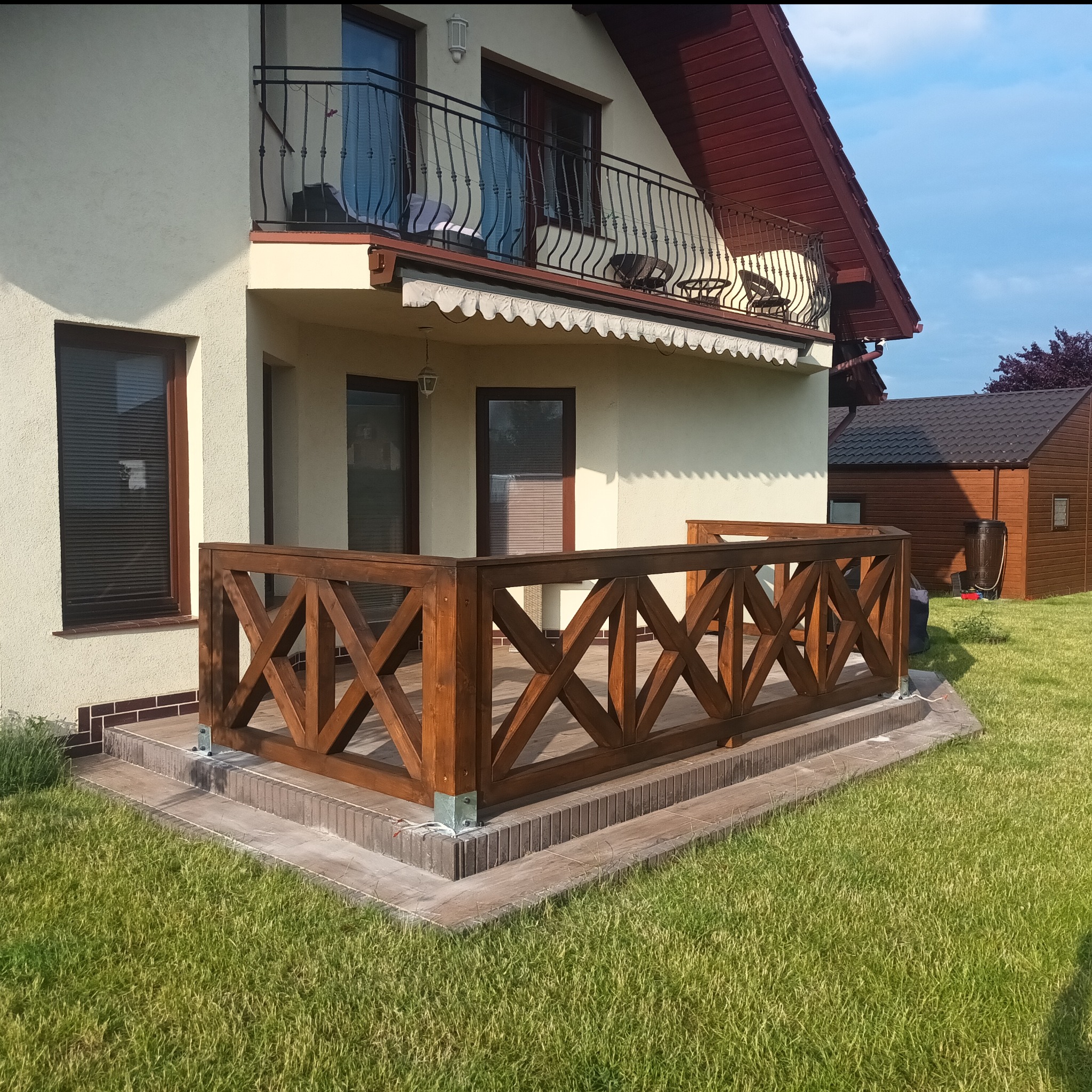 Drewniana balustrada tarasu w Barwicach z charakterystycznym wzorem krzyży, widok z poziomu trawnika na tle domu z balkonem i małym budynkiem gospodarczym.