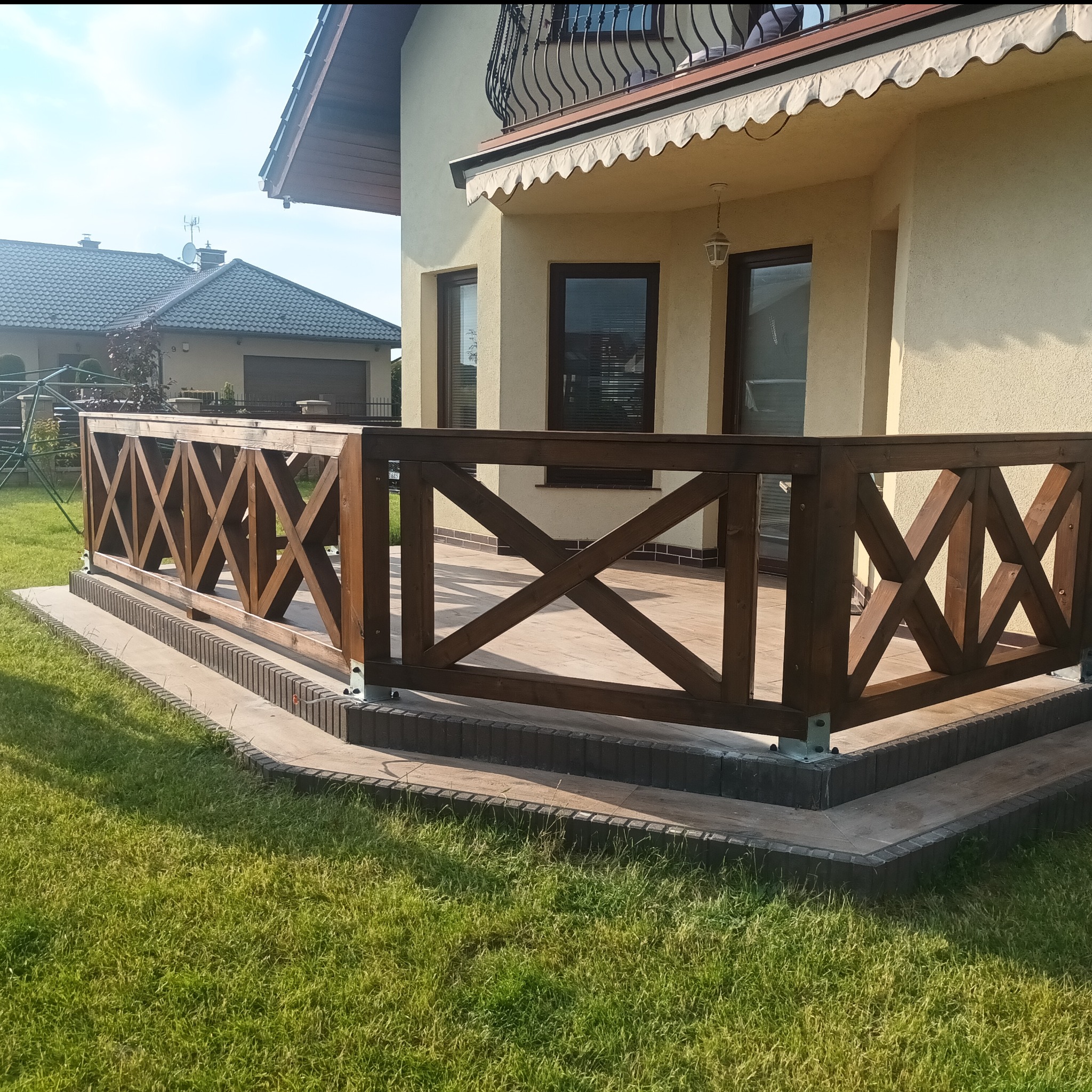 Ciemnobrązowa balustrada z krzyżowymi wzorami zabezpieczająca taras z jasnych płytek, widok z poziomu trawnika, w tle dom z beżową elewacją i ciemnym dachem.