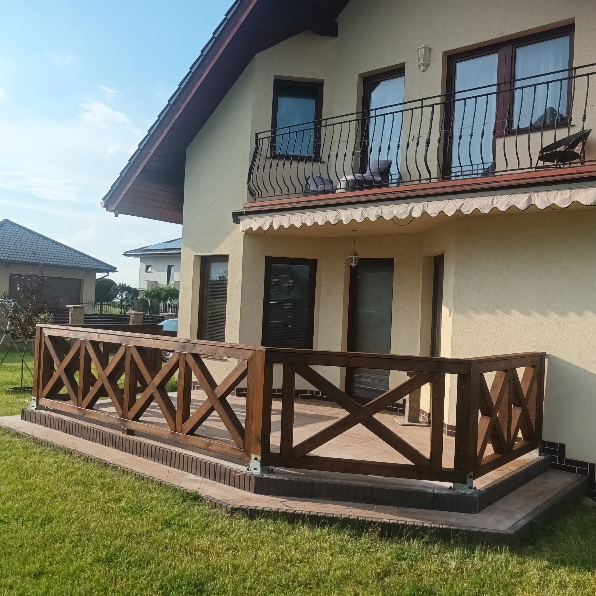 Zadaszony taras z drewnianą balustradą w kształcie krzyży, widok z poziomu trawnika, na drugim planie dom z balkonem.