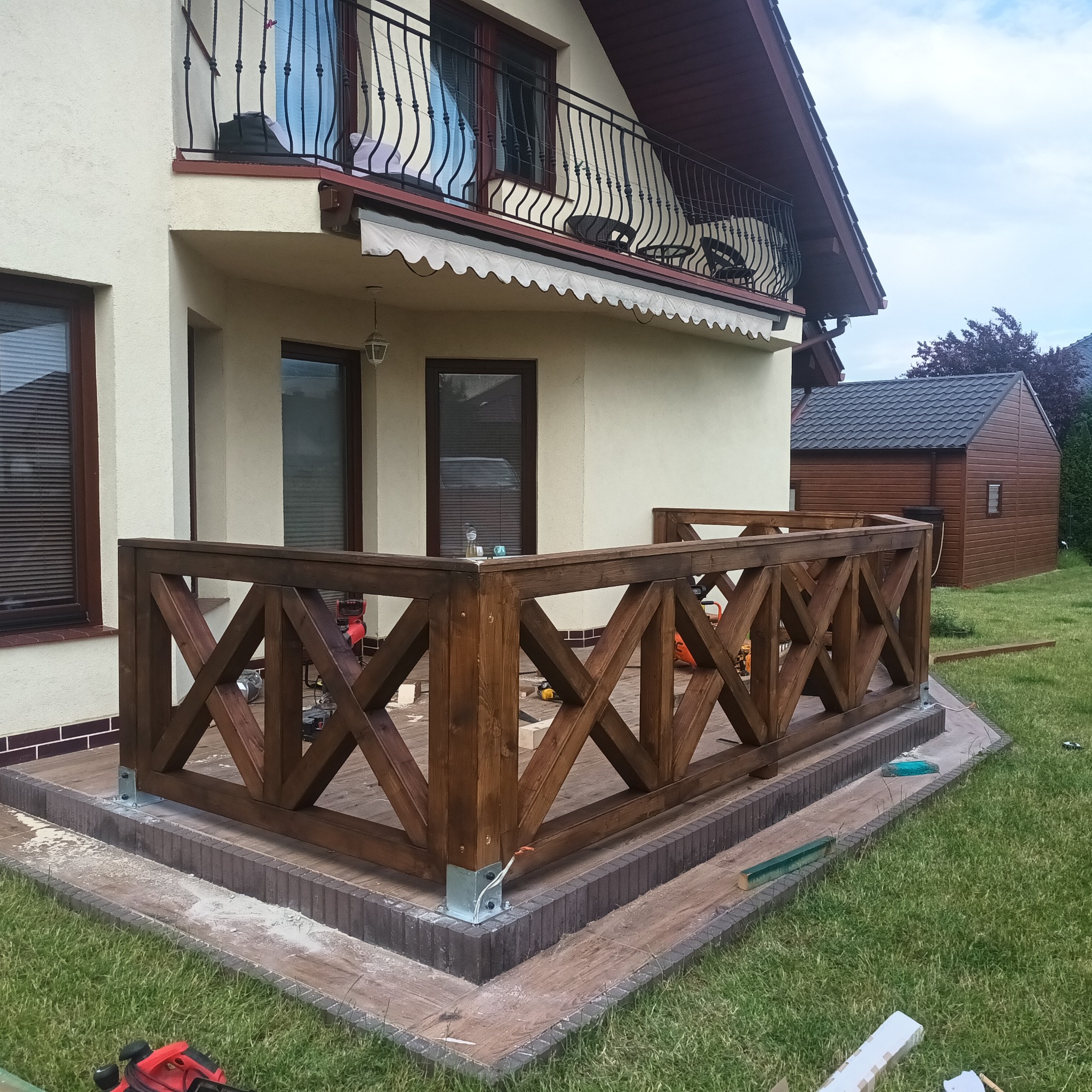 Nowo wybudowany, ciemnobrązowy, drewniany taras z krzyżowymi wzorami, częściowo umeblowany, z widocznymi narzędziami budowlanymi w tle, przylegający do jasnego domu z balkonem.