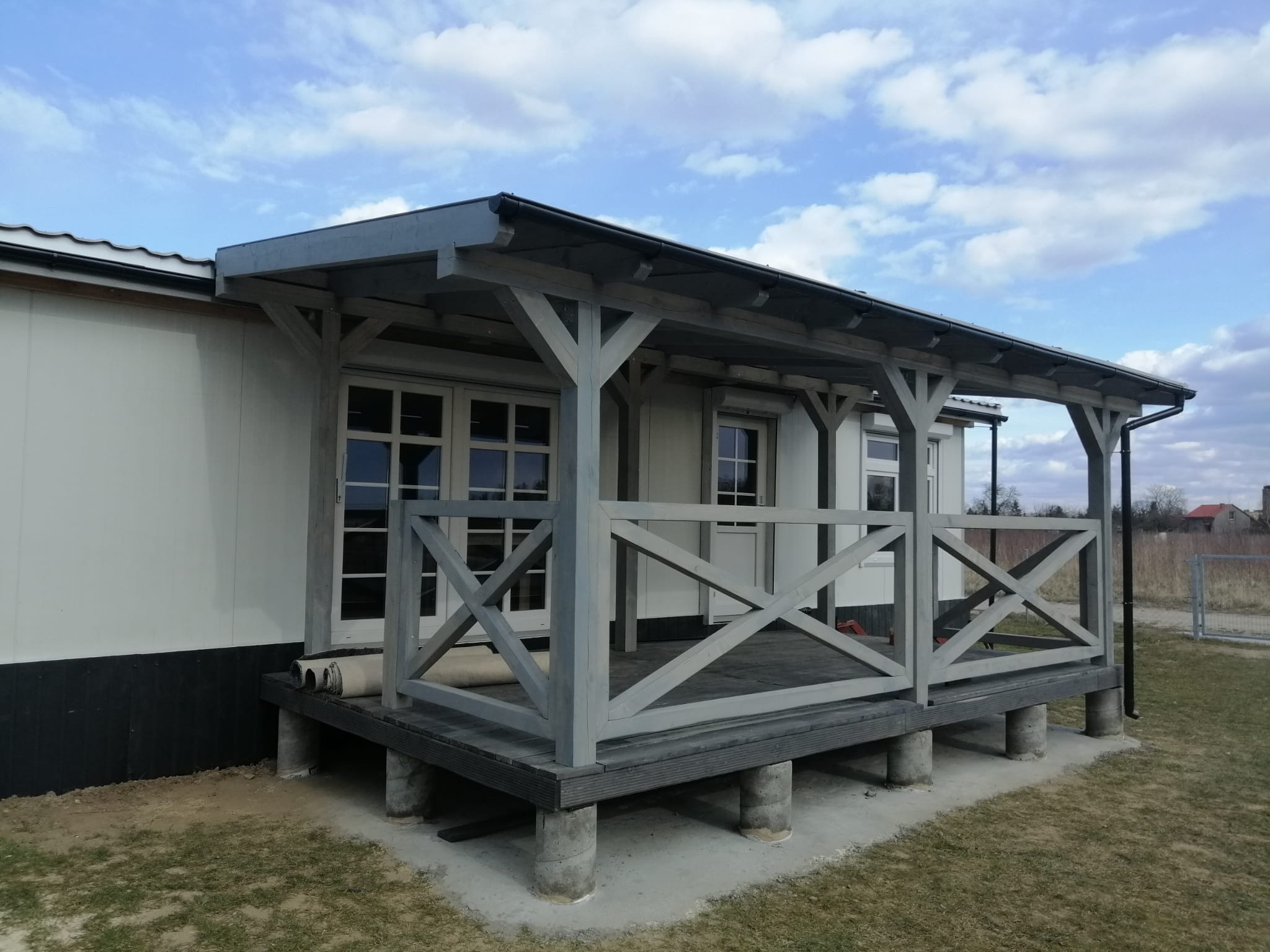 Zadaszony taras z drewnianą balustradą w kształcie krzyży, wsparty na betonowych słupach, przylegający do budynku z białymi ścianami i widocznymi rolkami materiału na podłodze.