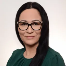 Portret kobiety w okularach z czarnymi włosami, ubranej w zieloną bluzkę, na jasnym tle.