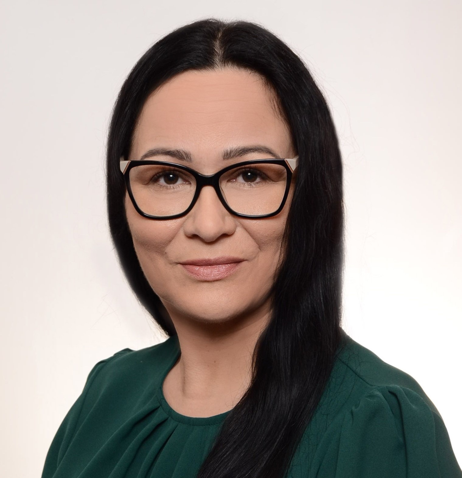 Portret kobiety w okularach z czarnymi włosami, ubranej w zieloną bluzkę, na jasnym tle.