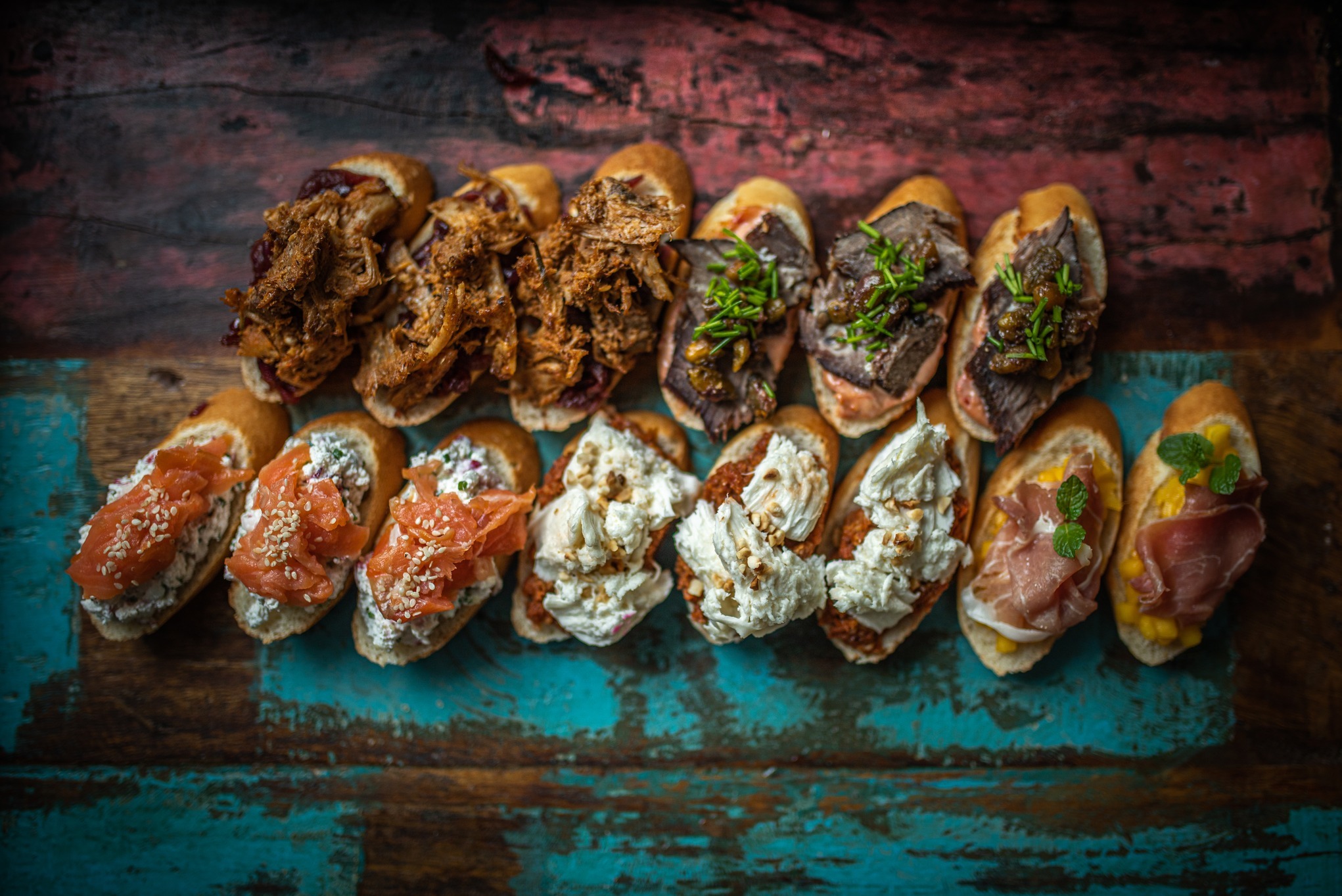 Różnorodne kanapki na rustykalnej desce: szarpana wieprzowina z sosem, rostbef z ziołami, łosoś z serkiem i sezamem, kremowy ser z orzechami, szynka parmeńska z melonem i miętą, widok z góry.